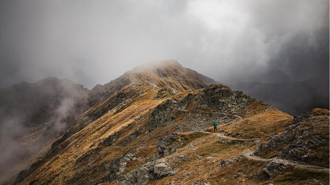 Episches Landschaftsbild: Ein  einzelner Trailrunner auf einem schmalen,  grasbewachsenen Grat, umgeben von  dramatischen, nebelverhangenen  Dolomitengipfeln bei kühler Morgenstimmung
