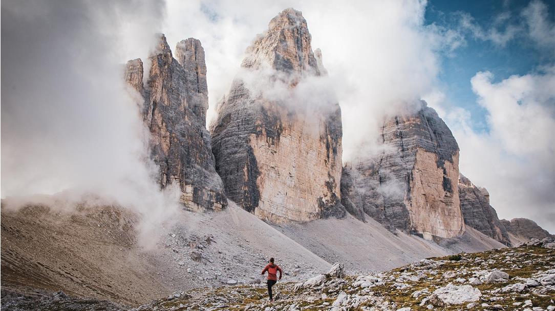 Trail runner in a red jacket on  a gravel path against the imposing,  cloud-shrouded backdrop of the Three  Peaks in the Dolomites.