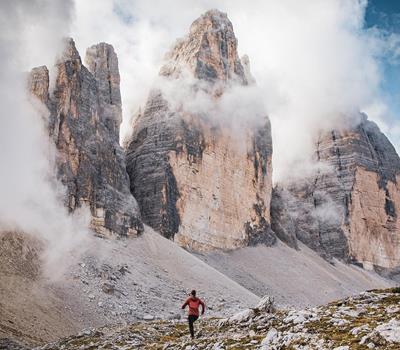 1 Trailrunner in roter Jacke auf einem  Schotterpfad vor der gewaltigen,  wolkenverhangenen Kulisse der Drei Zinnen in  den Dolomiten.