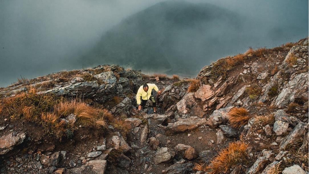 Close-up of a trail runner in a  bright yellow jacket and running shorts  while navigating a technical climb over  rough boulders and rocks in the steep Dolomites