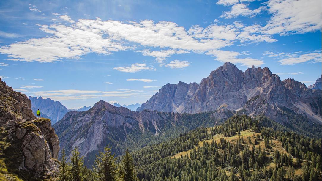 Spectacular panoramic view of  the high-alpine rock massifs of the  Dolomites. A trail runner in neonyellow clothing stands on an exposed  rocky ledge and enjoys the view. Far  below him lies a green valley.