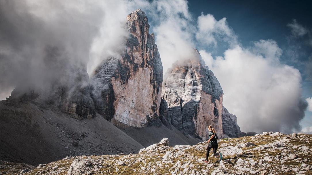 A female trail runner in  dynamic motion on an alpine gravel  path in front of the dramatically cloudshrouded north face of the Three Peaks  in the Dolomites.